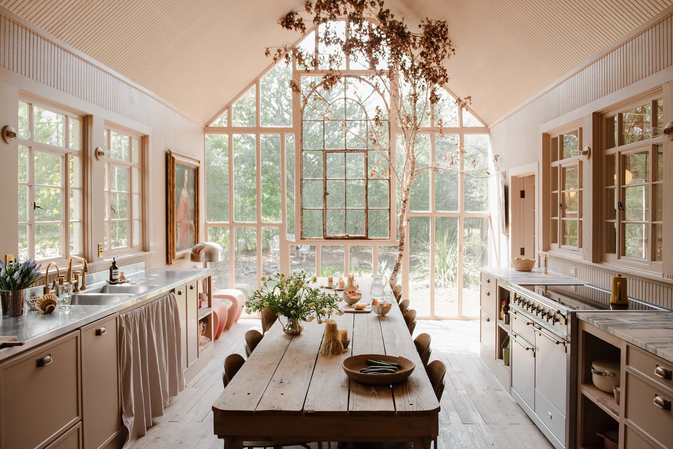 Long farmhouse table in a glasshouse kitchen with a dried-flower tree beneath an arched window